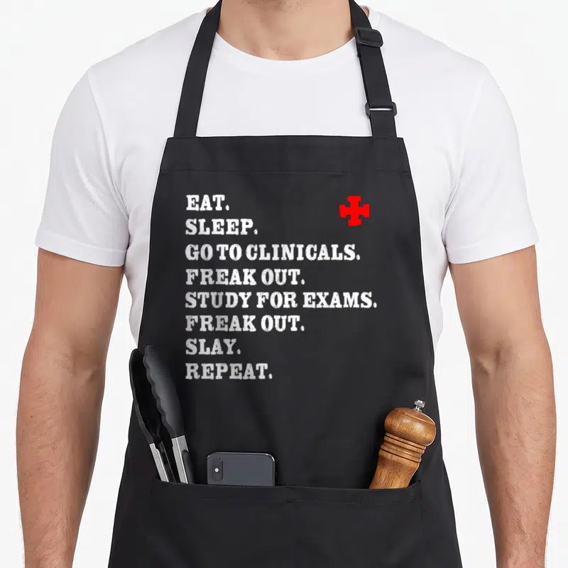 Slay nurse apron - White stacked text EAT SLEEP GO TO CLINICALS with red cross symbol — shown on a black cotton canvas apron against a white background