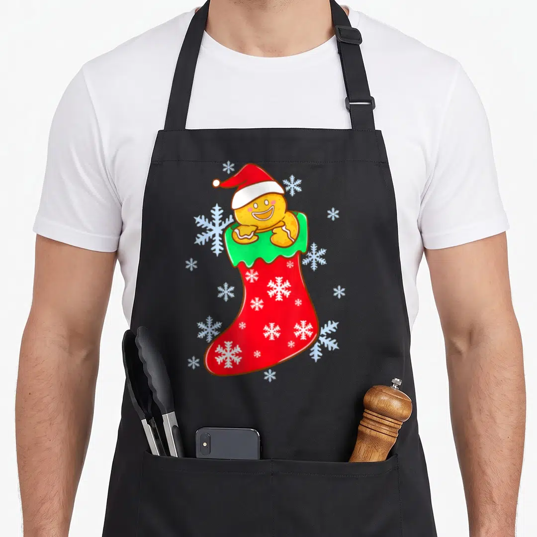 Stocking christmas gingerbread apron - Cartoon gingerbread man in santa hat inside red stocking with white snowflakes — shown on a black cotton canvas apron against a white background