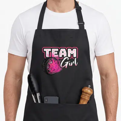 Burnout pregnancy by company apron - Car wheel with pink smoke and Team Girl text in white and pink — shown on a black cotton canvas apron against a white background