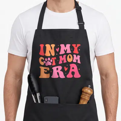 Era cat apron - Pink paw print replaces A in IN MY CAT MOM ERA text with hearts and sparkles — shown on a black cotton canvas apron against a white background