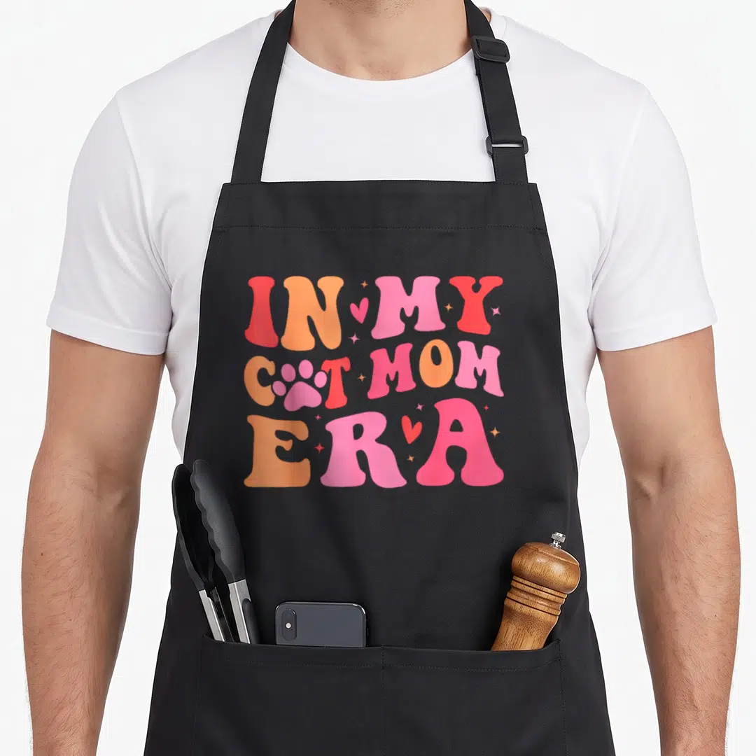 Era cat apron - Pink paw print replaces A in IN MY CAT MOM ERA text with hearts and sparkles — shown on a black cotton canvas apron against a white background