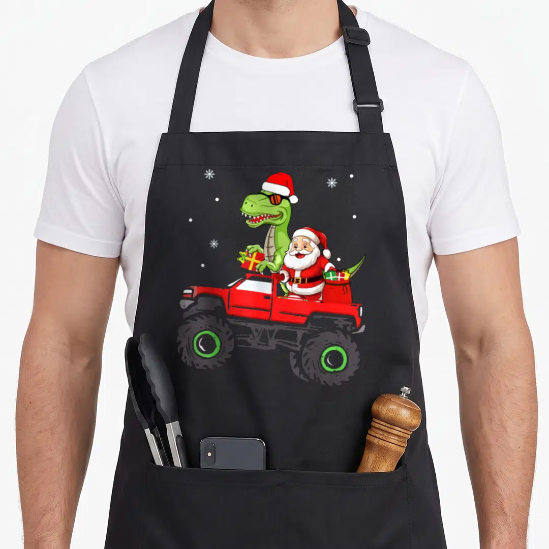Santa family christmas aprons - Green dinosaur in Santa hat and sunglasses riding red monster truck with Santa Claus — shown on a black cotton canvas apron against a white background