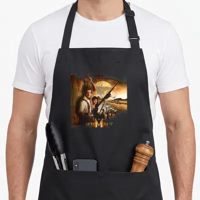 Rick mummy and me aprons - Man with rifle and rope beside woman, giant mummy silhouette, pyramids, helicopter — shown on a black cotton canvas apron against a white background