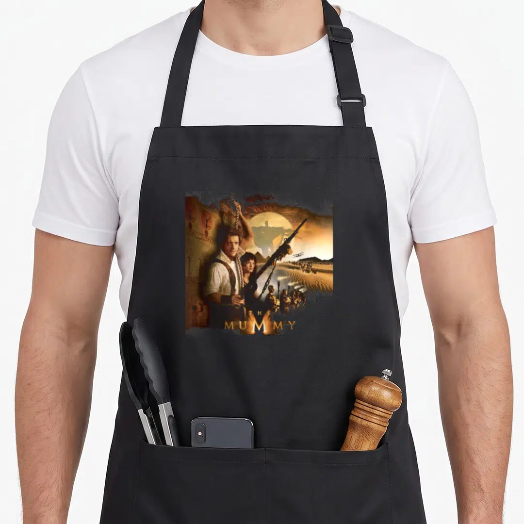 Rick mummy and me aprons - Man with rifle and rope beside woman, giant mummy silhouette, pyramids, helicopter — shown on a black cotton canvas apron against a white background