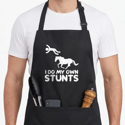 Stunt horse apron - White falling person and horse silhouette with I DO MY OWN STUNTS text — shown on a black cotton canvas apron against a white background