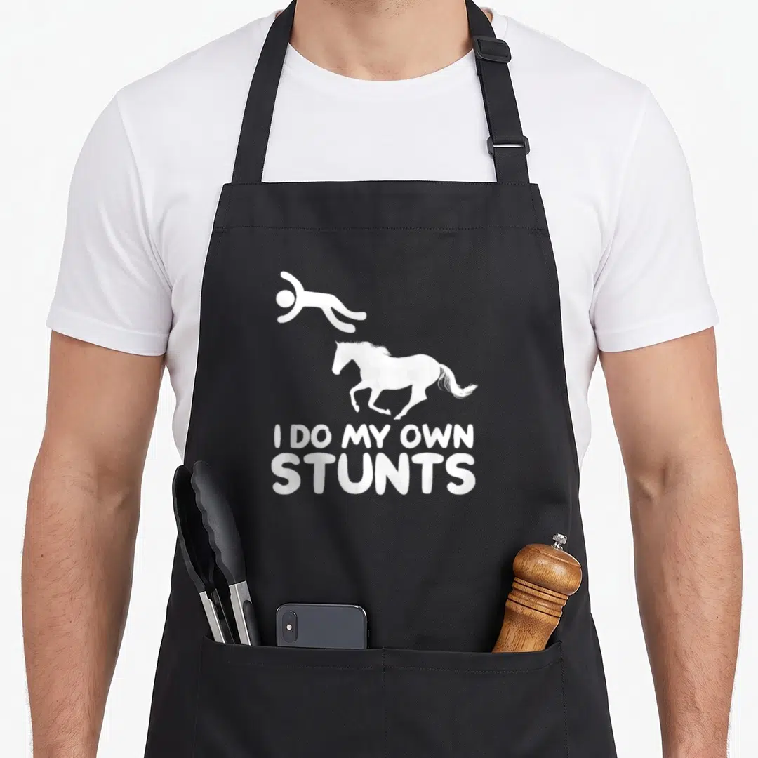 Stunt horse apron - White falling person and horse silhouette with I DO MY OWN STUNTS text — shown on a black cotton canvas apron against a white background