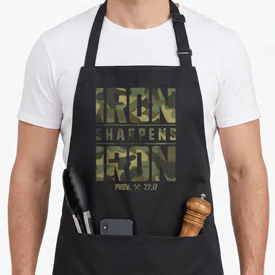 Iron camo apron - Olive green and tan camo text reading Iron Sharpens Iron Prov 27:17 with crossed hammers — shown on a black cotton canvas apron against a white background
