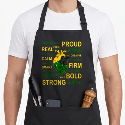 Jamaican creole apron - Anatomical heart in yellow green and black surrounded by words like PROUD BRAVE and STRONG — shown on a black cotton canvas apron against a white background