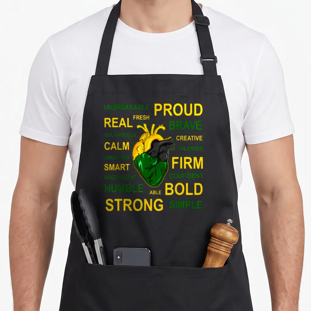 Jamaican creole apron - Anatomical heart in yellow green and black surrounded by words like PROUD BRAVE and STRONG — shown on a black cotton canvas apron against a white background
