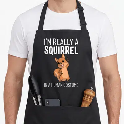 Squirrel halloween apron - Brown cartoon squirrel holding up fingers under white text reading I'M REALLY A SQUIRREL — shown on a black cotton canvas apron against a white background