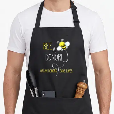 Kns bee apron - Cartoon bee with dotted trail and BEE A DONOR! text — shown on a black cotton canvas apron against a white background