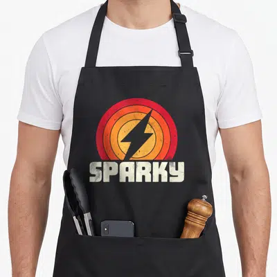 Funny sparky electricians apron - Grey lightning bolt in red orange yellow semi-circle with white SPARKY text below — shown on a black cotton canvas apron against a white background
