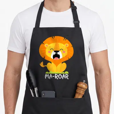 Roar passover apron - Cartoon lion sitting on grass with white MA-ROAR text below — shown on a black cotton canvas apron against a white background