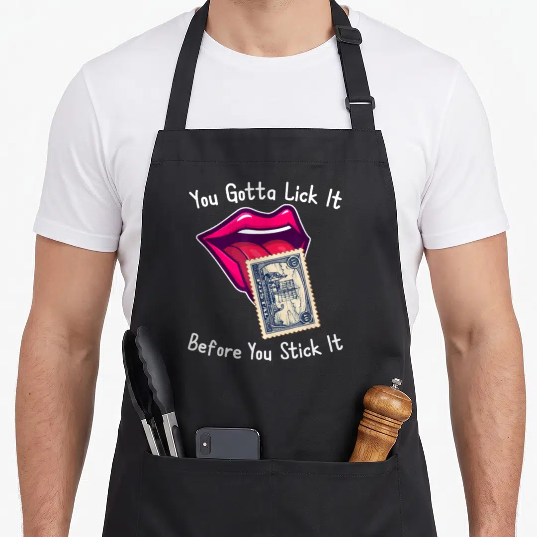 Lick joke apron - Cartoon mouth licking blue stamp with white text You Gotta Lick It Before You Stick It — shown on a black cotton canvas apron against a white background