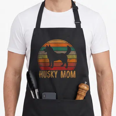 Pet husky apron - Grey husky silhouette over orange green yellow red grey striped sun with HUSKY MOM text — shown on a black cotton canvas apron against a white background