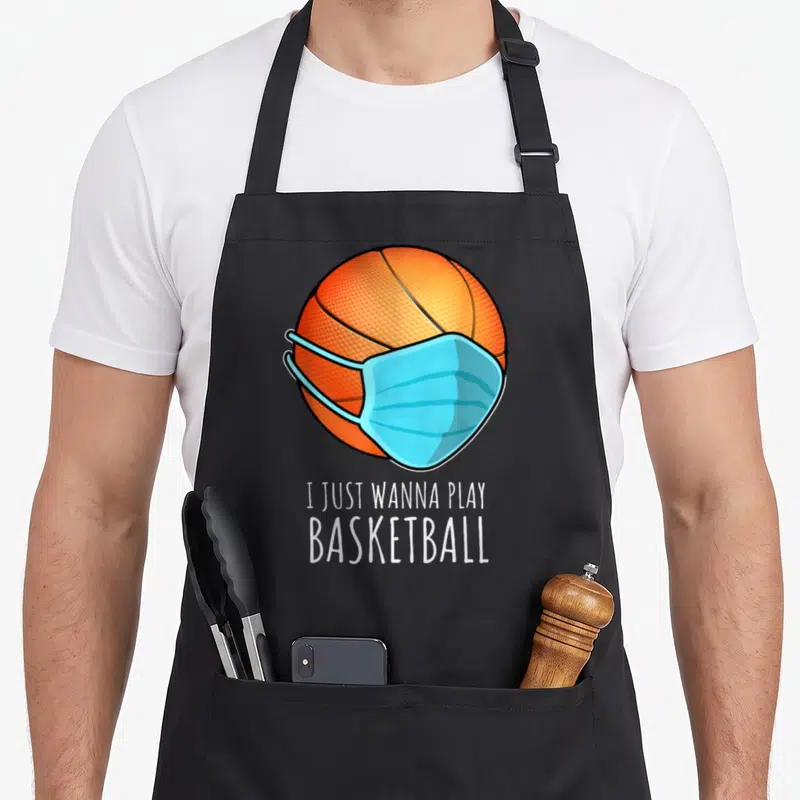 Play player basketball apron - Orange basketball wearing a light blue medical mask above white text reading I Just — shown on a black cotton canvas apron against a white background