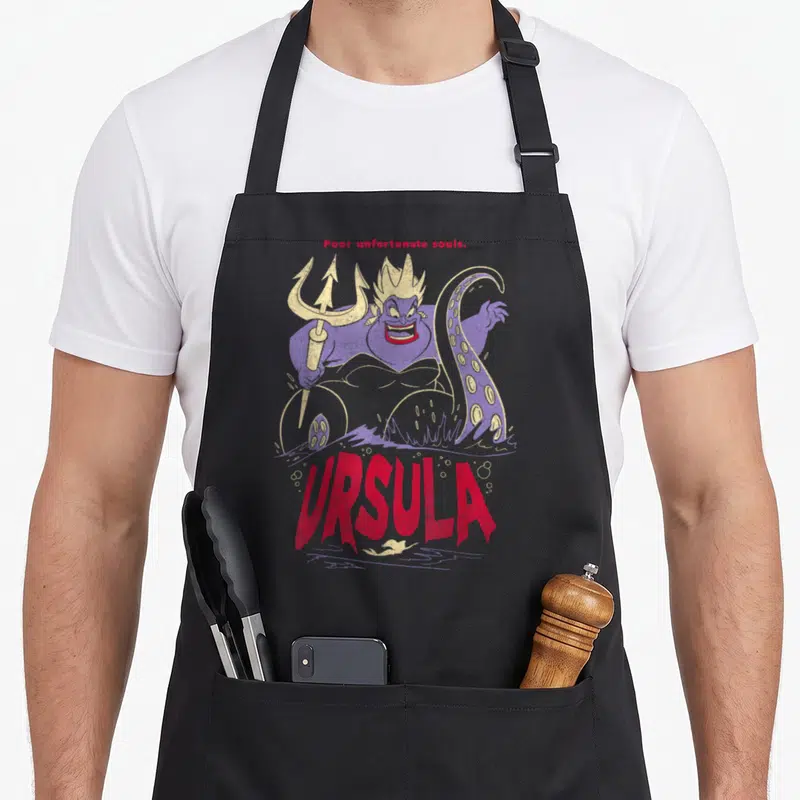 Ursula villain novelty apron - Purple Ursula holding gold trident with red text Poor unfortunate souls and URSULA — shown on a black cotton canvas apron against a white background
