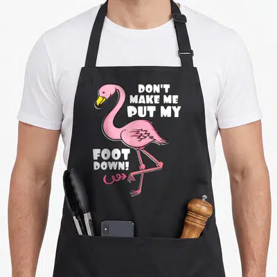 Foot bird pink flamingo apron - Cartoon pink flamingo with yellow beak and white text Don't Make Me Put My Foot Down — shown on a black cotton canvas apron against a white background
