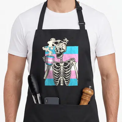 Transgender skeleton apron - Cartoon skeleton holding pride-striped mug over blue pink white flag background — shown on a black cotton canvas apron against a white background