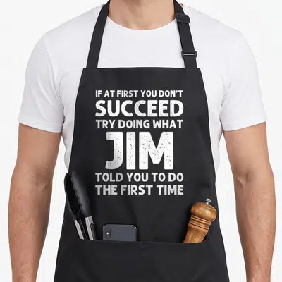 Crew husband apron - White stacked typography reading if at first you dont succeed try doing what jim told you to do — shown on a black cotton canvas apron against a white background