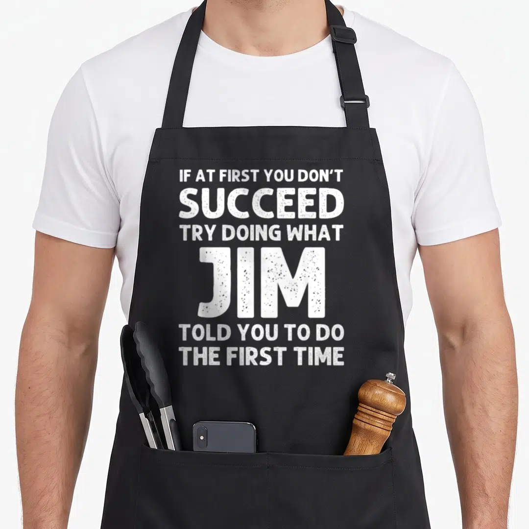 Crew husband apron - White stacked typography reading if at first you dont succeed try doing what jim told you to do — shown on a black cotton canvas apron against a white background