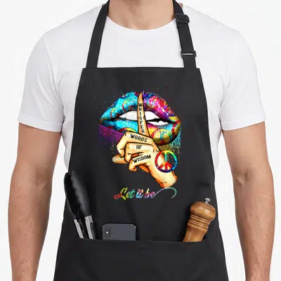 Whisper peace apron - Hand with upright finger over rainbow lips with peace signs and text whisper words of wisdom — shown on a black cotton canvas apron against a white background