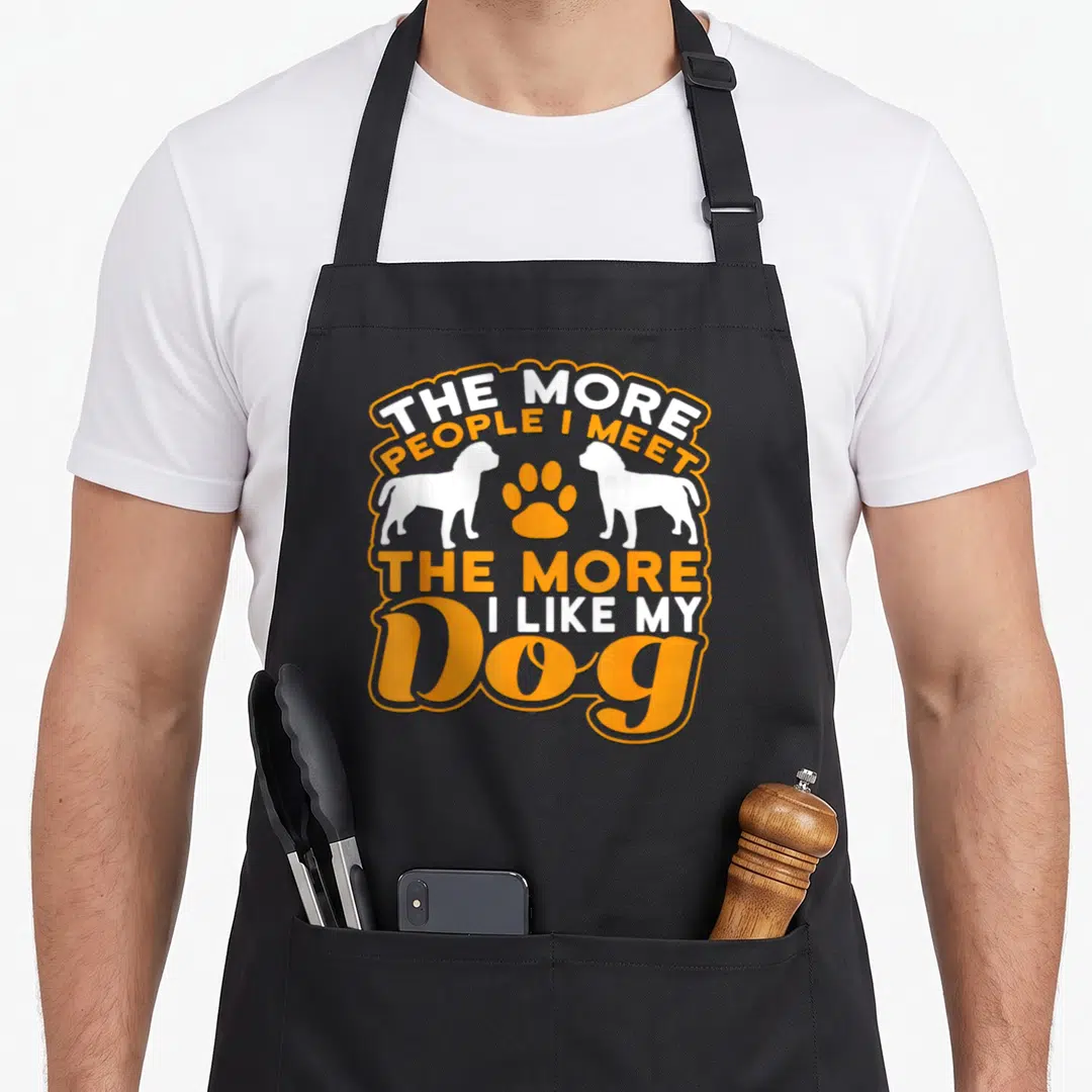 Funny meet dog apron - Orange and white text reading THE MORE PEOPLE I MEET THE MORE I LIKE MY DOG flanking two dog — shown on a black cotton canvas apron against a white background