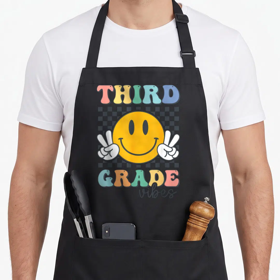 Smile grade vibes apron - Third Grade vibes text with yellow smiley face holding white peace signs on checkerboard — shown on a black cotton canvas apron against a white background