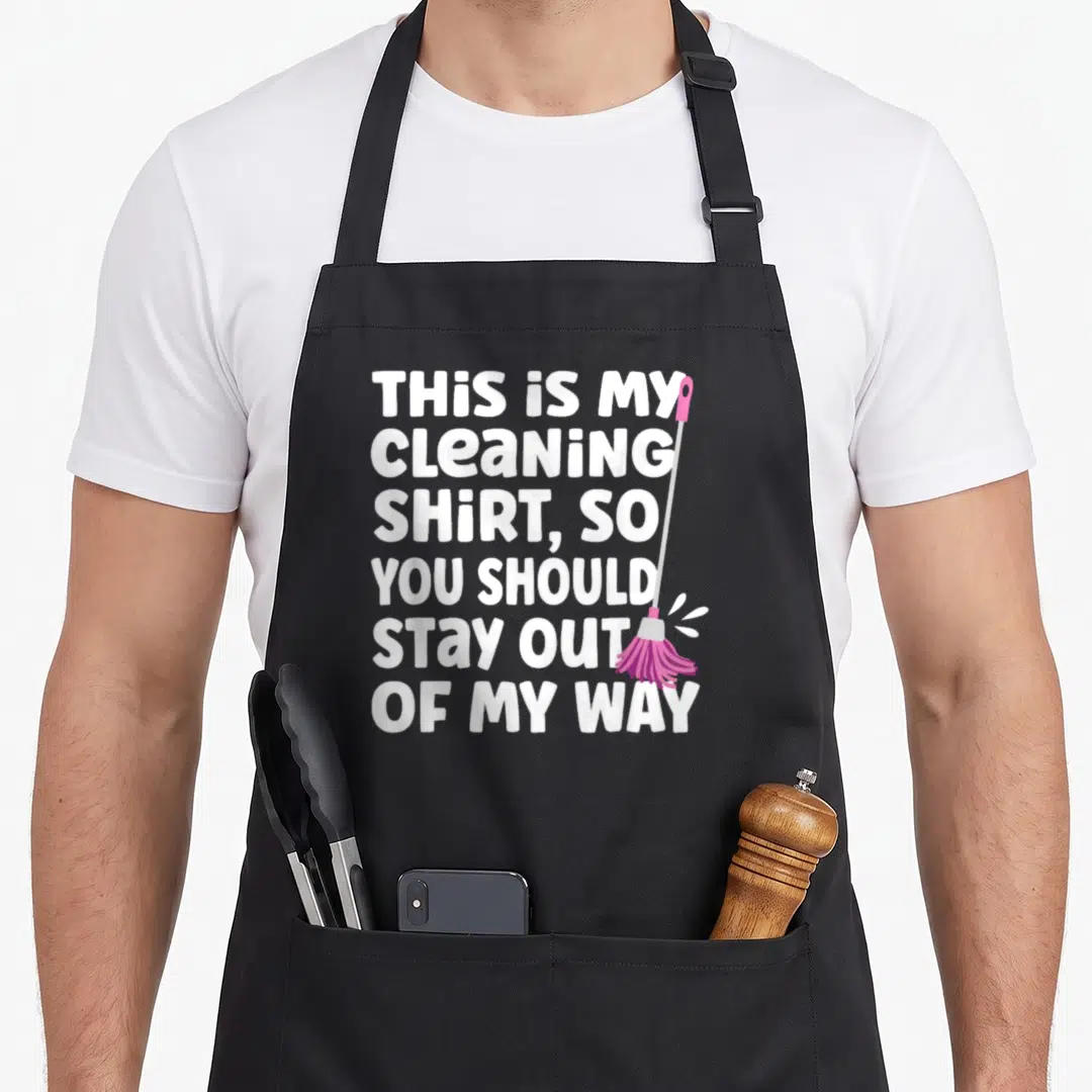 Freak cleaning apron - White text reading THIS IS MY CLEANING SHIRT beside a pink and grey mop — shown on a black cotton canvas apron against a white background