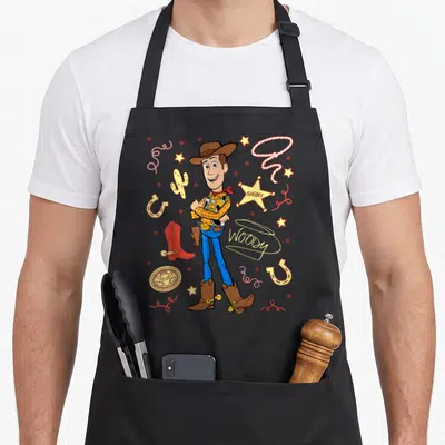 Sheriff western apron - Cartoon Woody standing among yellow stars, red lasso, and brown boots with SHERIFF text — shown on a black cotton canvas apron against a white background