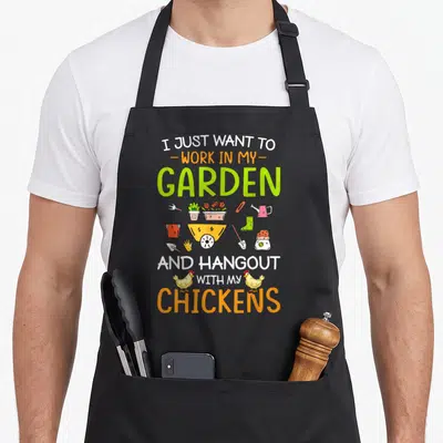 Hangout chicken apron - Cartoon gardening tools and two yellow chickens surround stacked text about working in a garden — shown on a black cotton canvas apron against a white background