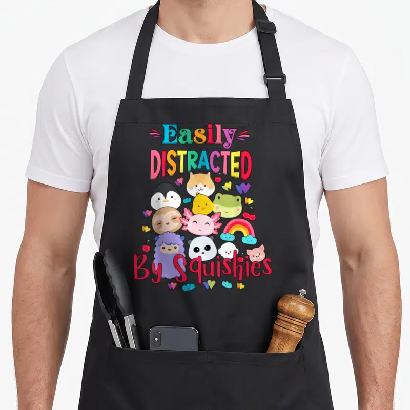 Squishy animal apron - Cartoon animals rainbow hearts cherry text easily distracted by squishies — shown on a black cotton canvas apron against a white background