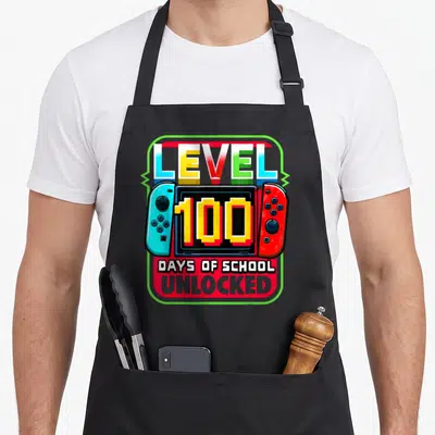 Level school game aprons - Cartoon game console with blue and red controllers displaying 100 DAYS OF SCHOOL UNLOCKED — shown on a black cotton canvas apron against a white background