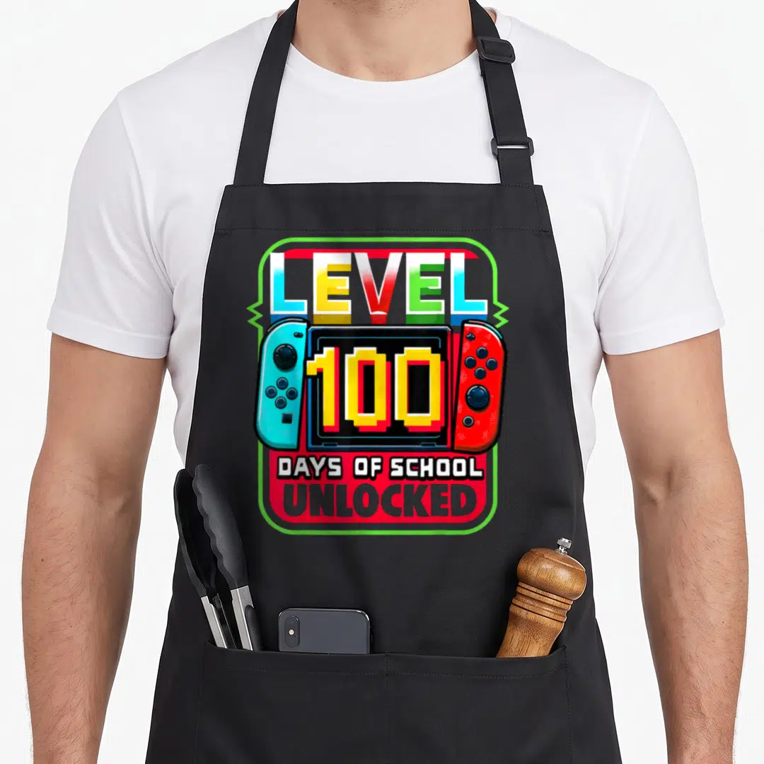 Level school game aprons - Cartoon game console with blue and red controllers displaying 100 DAYS OF SCHOOL UNLOCKED — shown on a black cotton canvas apron against a white background