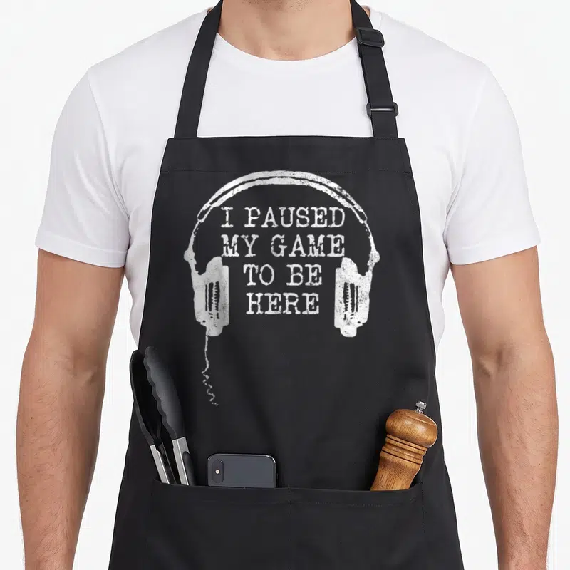 Gamer paused gaming apron - Distressed white headphones arching over stacked text reading I Paused My Game To Be Here — shown on a black cotton canvas apron against a white background