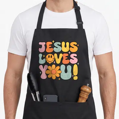 Church funny christian aprons - Retro JESUS LOVES YOU text with smiley face and flower in pink orange yellow blue — shown on a black cotton canvas apron against a white background