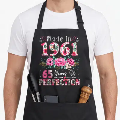 65th birthday vintage floral apron - Watercolor pink roses and leaves surrounding Made in 1961 and 65 Years — shown on a black cotton canvas apron against a white background