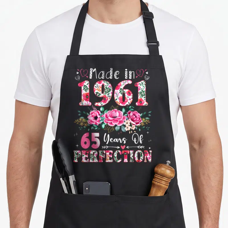 65th birthday vintage floral apron - Watercolor pink roses and leaves surrounding Made in 1961 and 65 Years — shown on a black cotton canvas apron against a white background