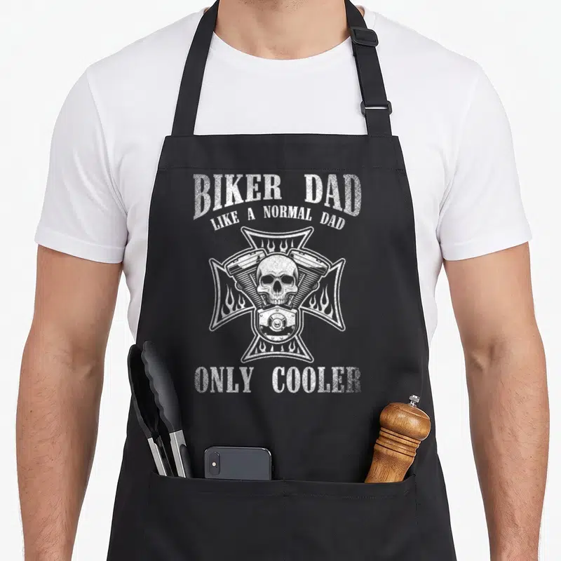 Cooler biker apron - White distressed skull over motorcycle engine inside a cross with flame details and arched text — shown on a black cotton canvas apron against a white background