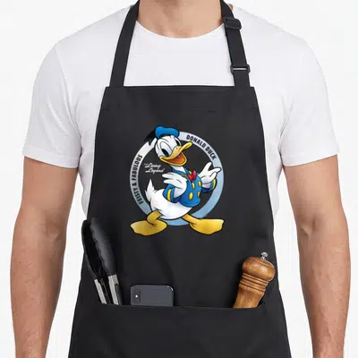90th anniversary apron - Donald Duck cartoon figure inside blue circle with Feisty and Fabulous text — shown on a black cotton canvas apron against a white background