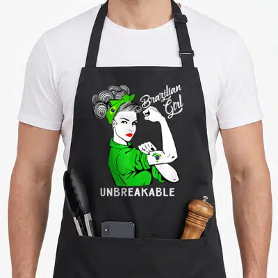 Brazilian heritage aprons - Cartoon woman flexing arm with Brazilian Girl UNBREAKABLE text and flag details — shown on a black cotton canvas apron against a white background