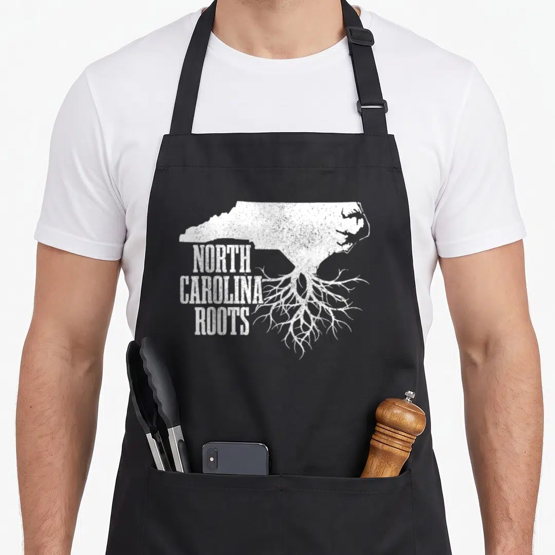 North patriotic apron - Distressed white NC map silhouette with roots and stacked text — shown on a black cotton canvas apron against a white background