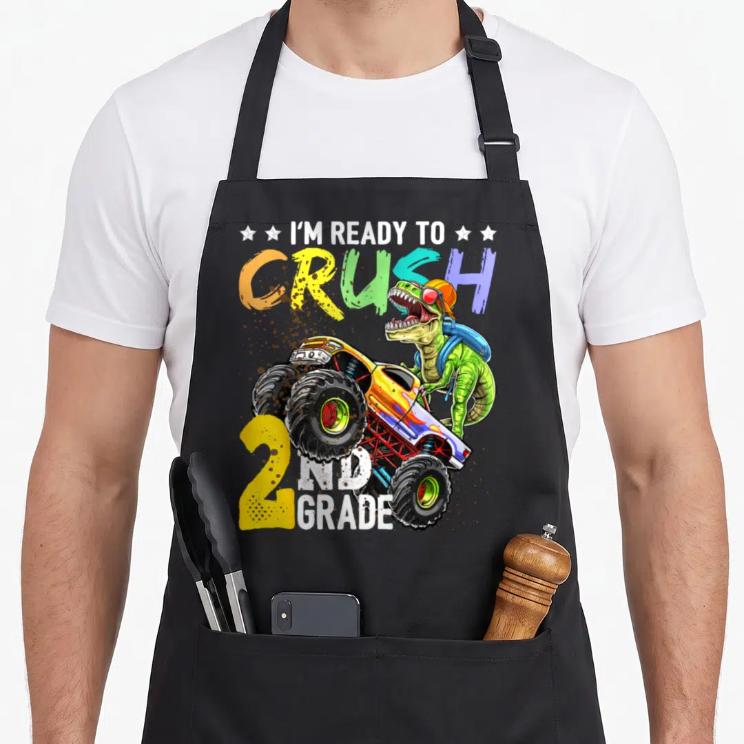Crush 2nd truck apron - T-Rex in hat glasses backpack next to monster truck with rainbow text and stars — shown on a black cotton canvas apron against a white background