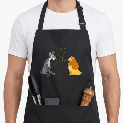 Bella lady and the tramp apron - Two cartoon dogs facing each other with a yellow string heart connecting their mouths — shown on a black cotton canvas apron against a white background