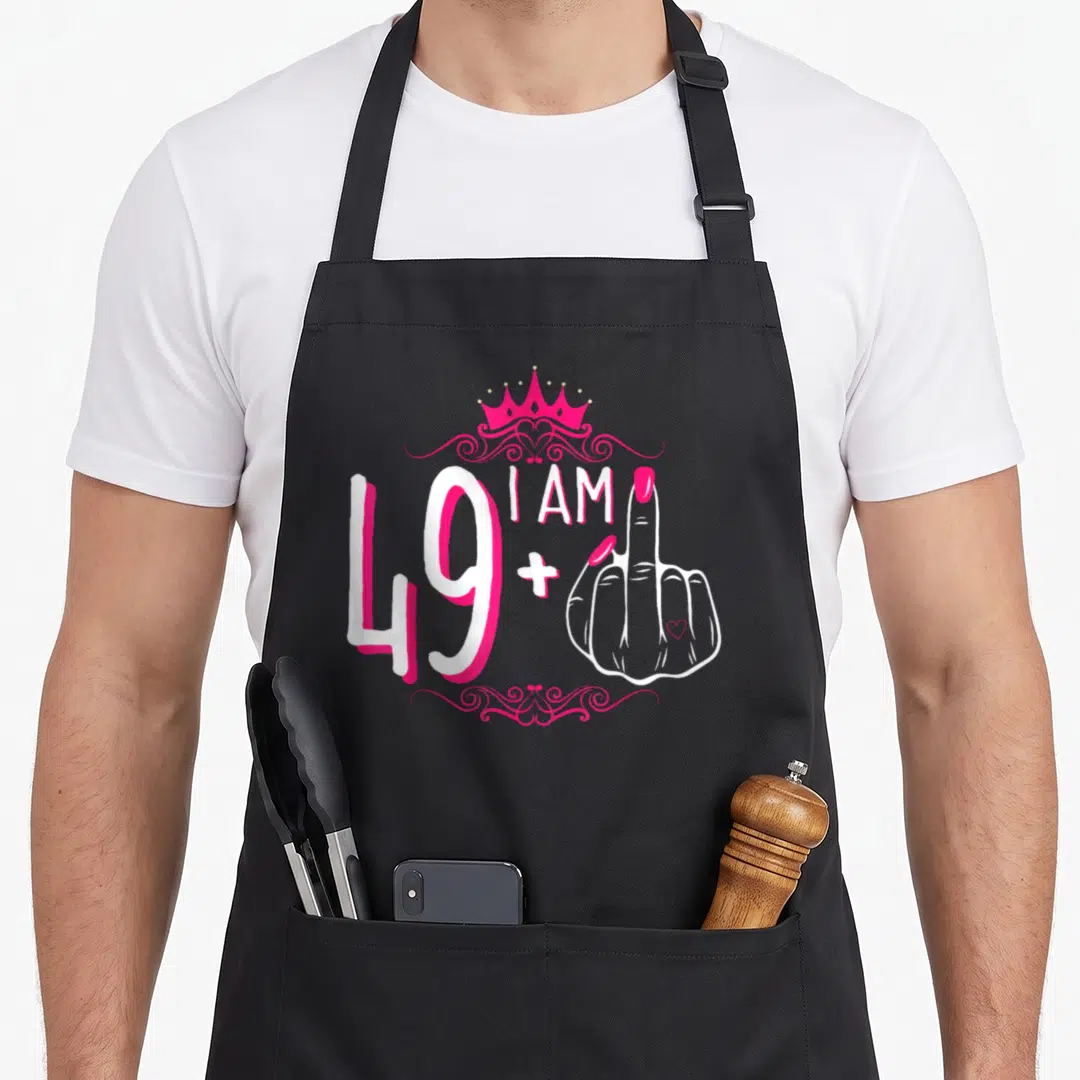 Finger 49th birthday apron - Cartoon middle finger hand with pink crown and I AM 49+ text — shown on a black cotton canvas apron against a white background