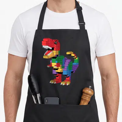 Dinosaur building apron - Cartoon Tyrannosaurus Rex built from red yellow green blue and purple interlocking plastic — shown on a black cotton canvas apron against a white background