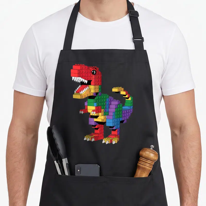 Dinosaur building apron - Cartoon Tyrannosaurus Rex built from red yellow green blue and purple interlocking plastic — shown on a black cotton canvas apron against a white background