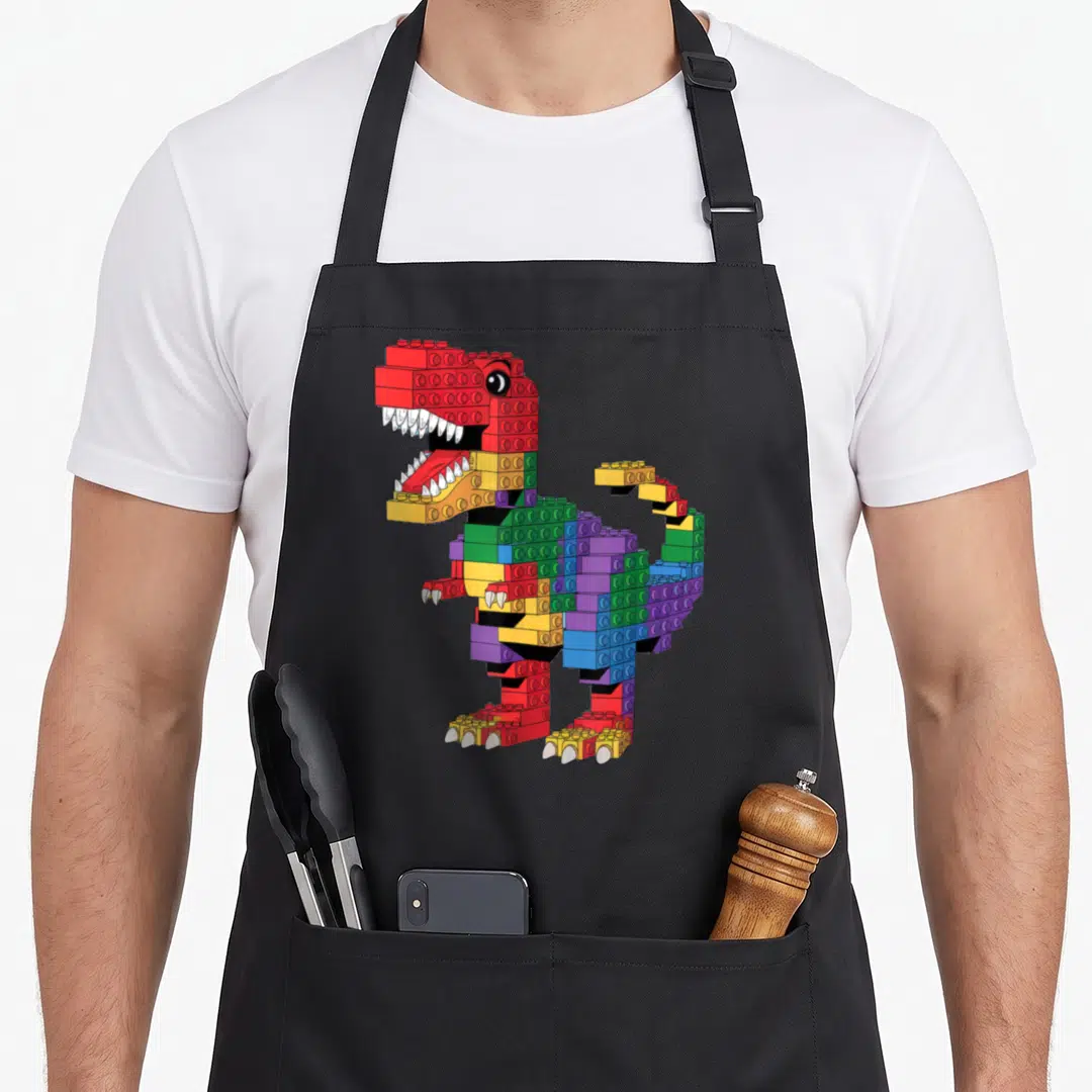 Dinosaur building apron - Cartoon Tyrannosaurus Rex built from red yellow green blue and purple interlocking plastic — shown on a black cotton canvas apron against a white background