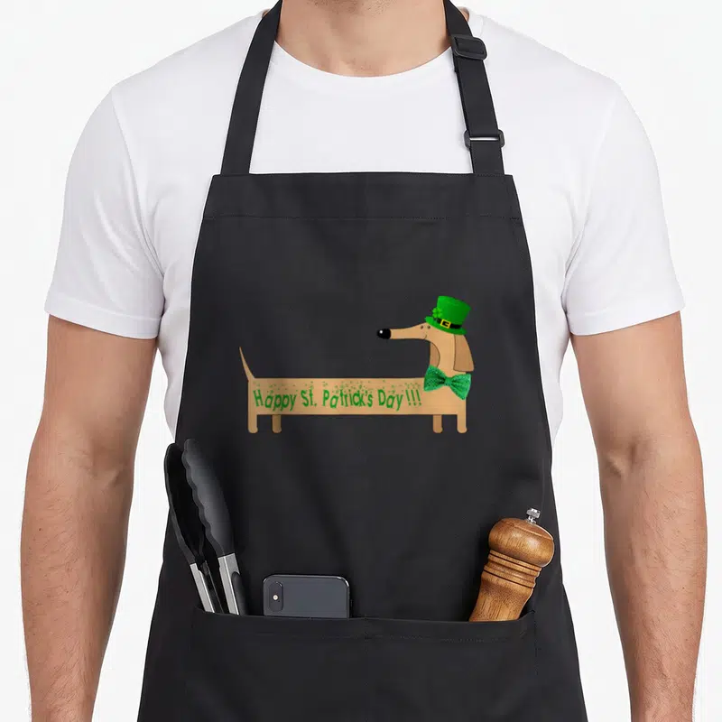 Saint wiener apron - Brown dachshund in profile wearing green top hat and bow tie with holiday text on side — shown on a black cotton canvas apron against a white background