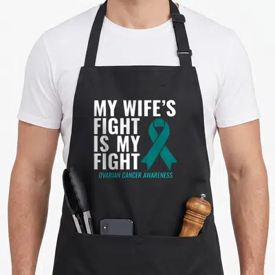Fight ovarian wife apron - Stacked white text and teal ribbon reading My Wife's Fight Is My Fight Ovarian Cancer — shown on a black cotton canvas apron against a white background